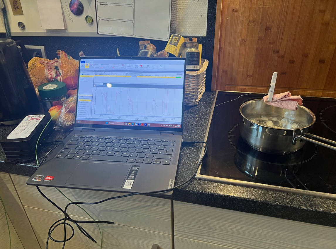 Auf einem Küchenherd wird ein Topf mit Eiswasser zum Kochen gebracht. Mit einer Wäscheklammer sind zwei Thermoelemente eines Widerstandsthermometers fixiert. Ein angeschlossener Laptop zeichnet die Daten auf.